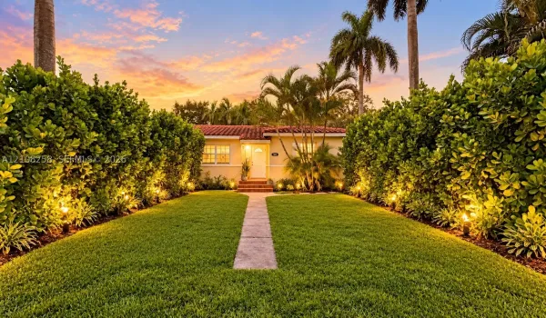 Exterior facade featuring a tile roof, straight concrete walkway, manicured lawn, dense perimeter hedge landscaping, and palm trees