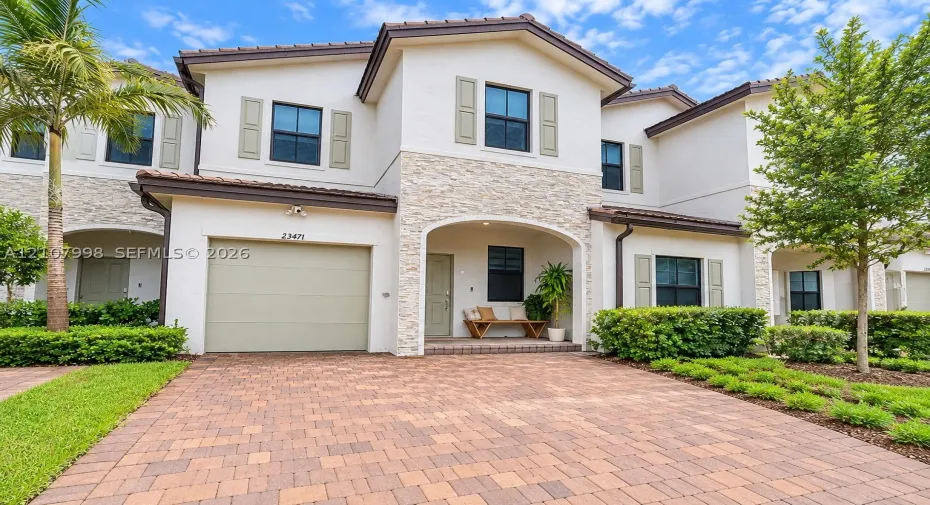 Two-story exterior featuring light-toned stucco, decorative stone veneer accents, a paver driveway, an arched covered entryway, and shutter-framed windows