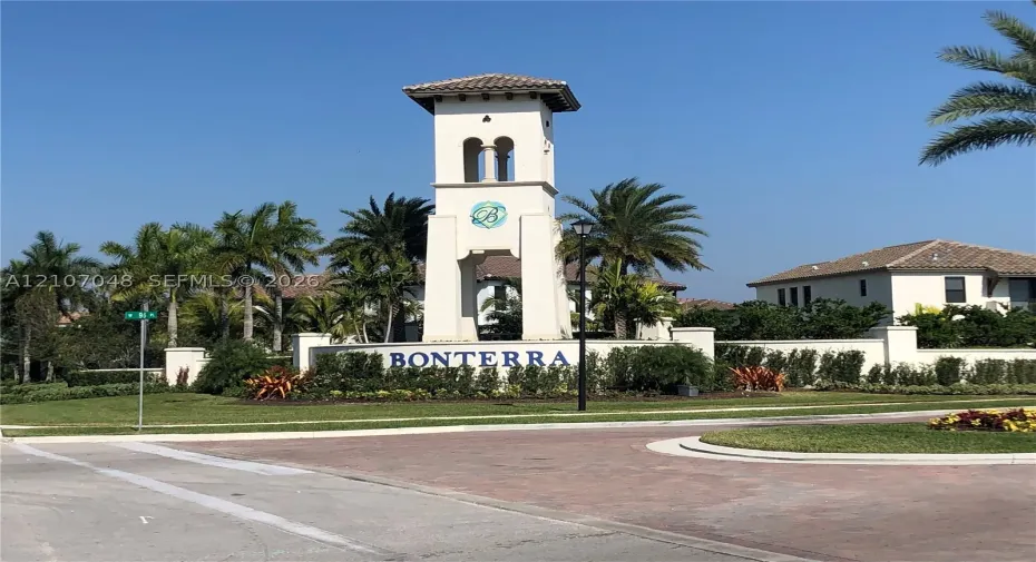 Community entrance featuring a landmark bell tower, stucco perimeter wall, and manicured landscaping with mature palm trees