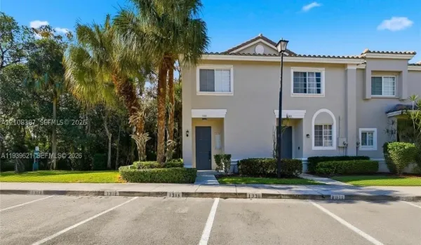 Two-story exterior featuring a neutral stucco facade, tile roofing, decorative arched window detailing, and integrated landscape hedging