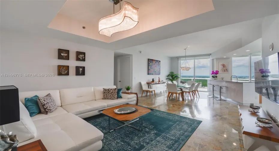 Open-concept living space featuring high tray ceilings with an oversized modern chandelier, polished stone flooring, a built-in kitchen peninsula with stone countertops, and floor-to-ceiling windows