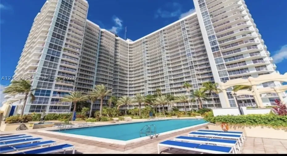 High-rise building exterior featuring a multi-story facade with wraparound balconies, accompanied by an outdoor swimming pool, paved sun deck, and mature palm trees