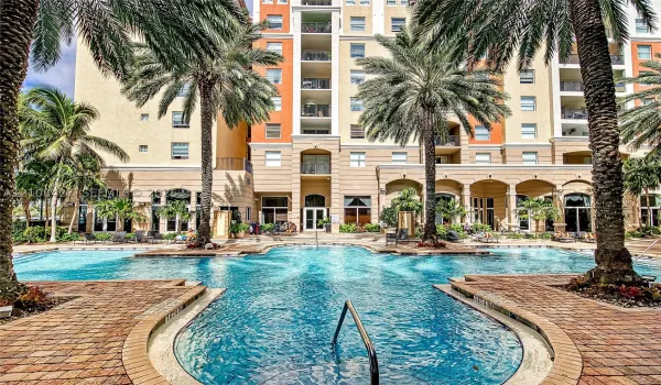 Resort-style swimming pool featuring a brick-paver deck, arched architectural detailing, mature palm trees, and multi-story residential facade