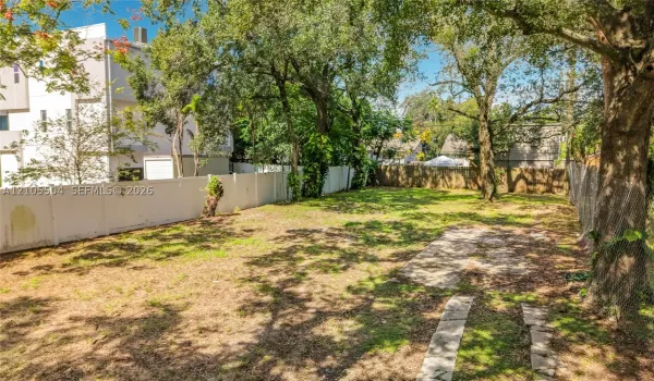 Expansive backyard featuring a white privacy fence, mature trees, and natural ground cover