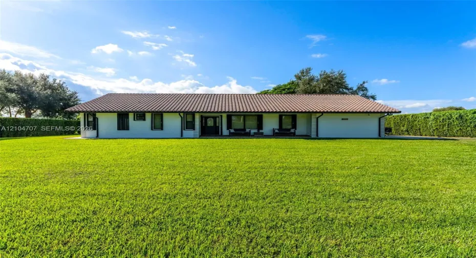 Single-story residence featuring a low-pitched tiled roof, light-toned exterior finish, and an expansive manicured lawn bordered by mature hedge fencing
