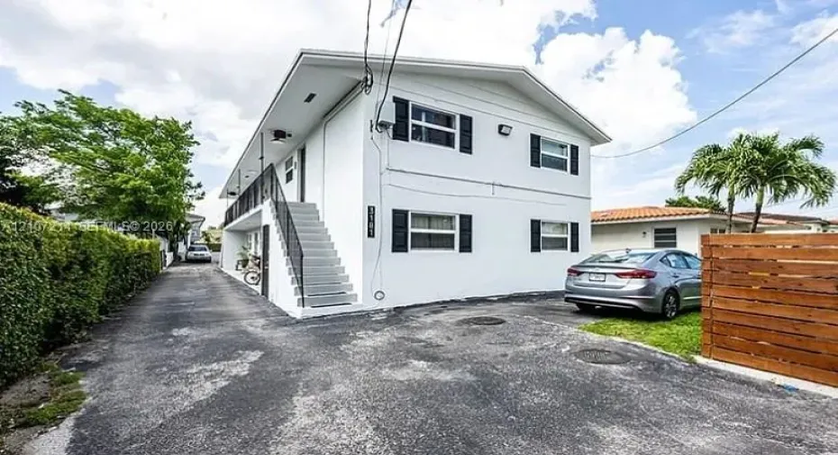 Two-story exterior featuring a white stucco facade, black shutters, external concrete staircase with metal railing, and asphalt paving