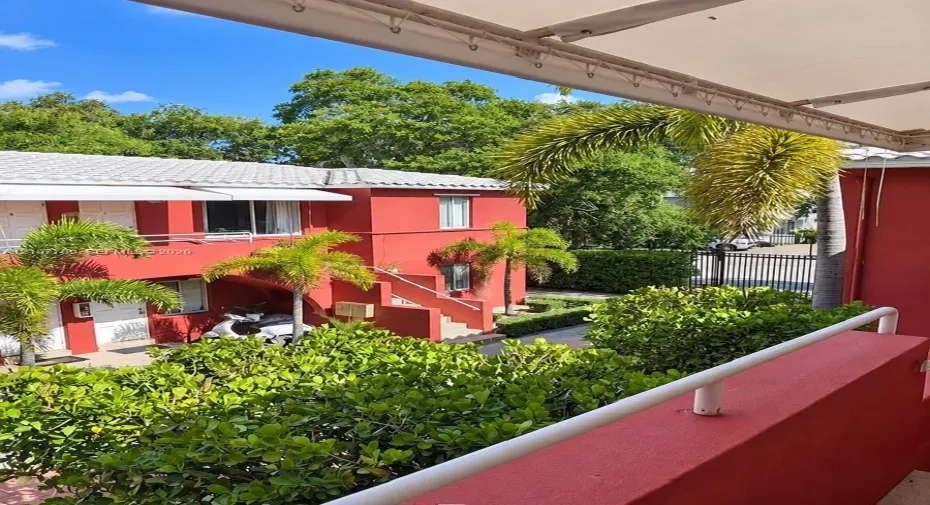 Balcony featuring a metal railing and retractable awning overlooking grounds with mature landscaping, palm trees, and exterior stairs