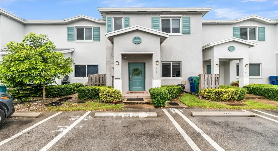 Two-story stucco exterior featuring gabled rooflines, teal window shutters, and assigned parking spaces