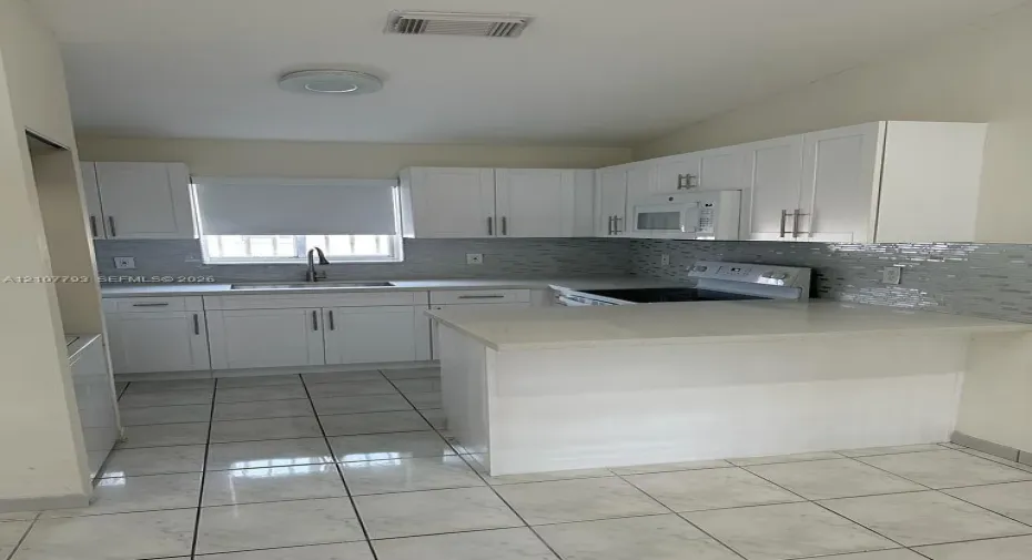 Kitchen featuring white shaker-style cabinetry, light-toned stone countertops, a mosaic tile backsplash, a peninsula, and tile flooring