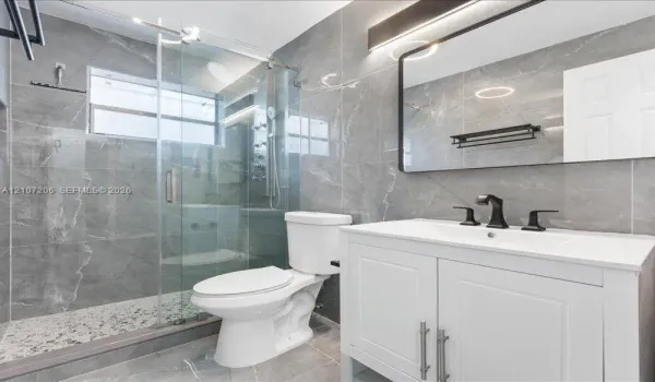 Full-height gray tiled walls, glass-enclosed shower with pebble-stone flooring, white vanity cabinet with a neutral countertop, matte black fixtures, and a modern rectangular mirror with integrated lighting