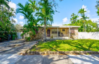Single-story residence featuring a stone-veneer facade, front porch with decorative metal railing, concrete driveway, and established palm landscaping enclosed by vinyl fencing