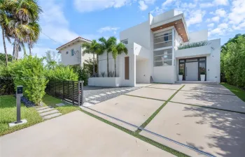 Modern architectural exterior featuring a multi-level white facade, geometric concrete paver driveway, full-height glass window wall, and integrated wood-finish ceiling accents