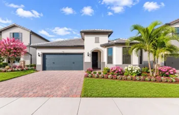 Stucco exterior with a tile roof, paver driveway, two-car garage, and manicured landscaping featuring flowering shrubs and mature palm trees