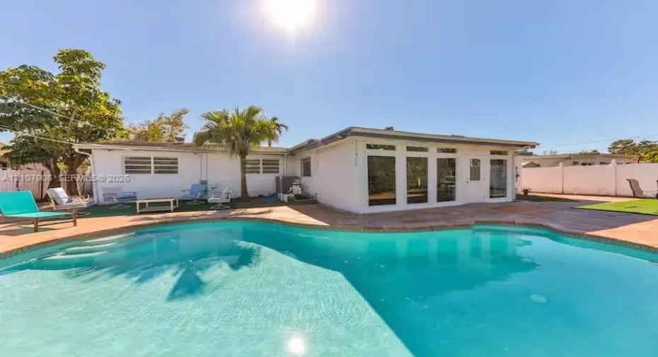 In-ground pool with stone paver surround, expansive patio space, white exterior siding, and custom glass-panel entry door