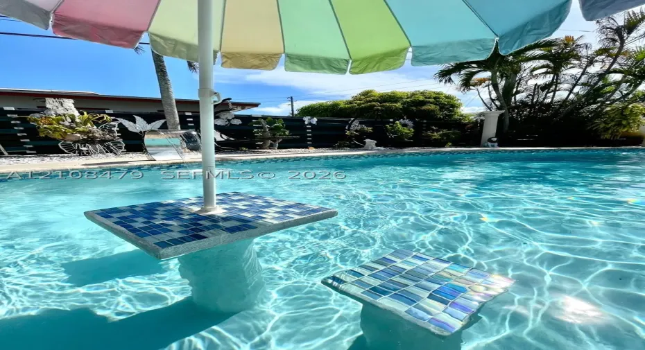 In-ground swimming pool featuring a tiled, built-in pedestal table and submerged seating with iridescent mosaic finish