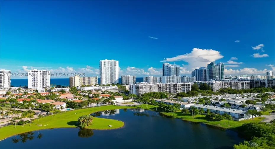 Aerial perspective featuring a central freshwater lake, manicured green space, mature palm trees, and surrounding coastal cityscape