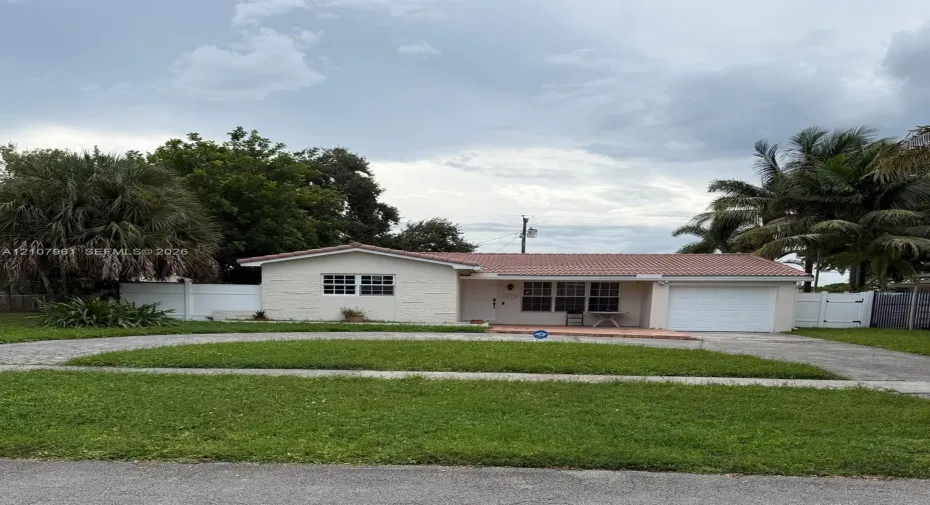 Single-story residence featuring a terracotta-tiled roof, textured stucco facade, attached garage, paved circular driveway, and white vinyl perimeter fencing