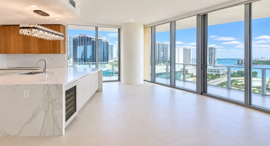 Open-concept kitchen featuring an expansive waterfall-edge island, stone-surfaced countertops, custom wood cabinetry, integrated wine refrigerator, and floor-to-ceiling glass windows