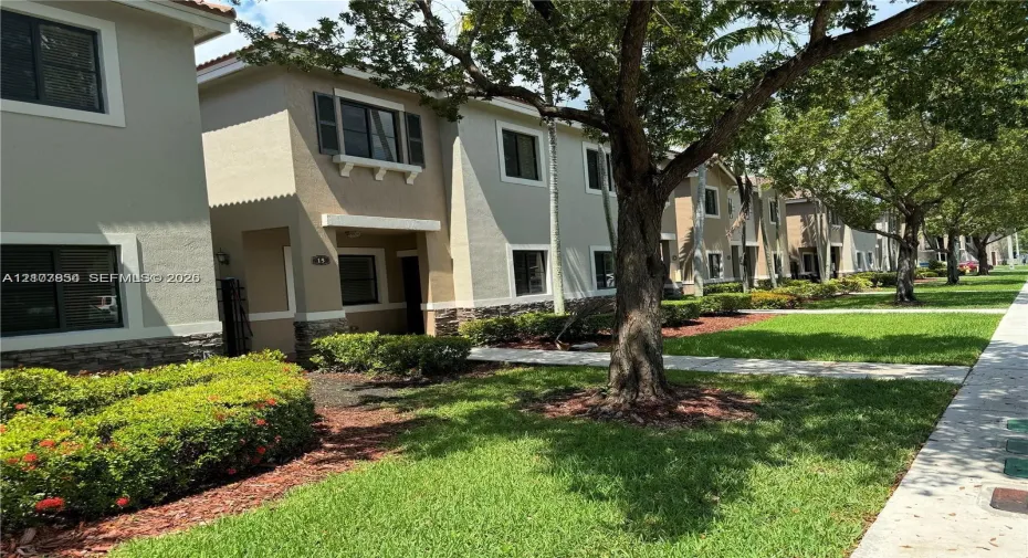 Two-story exterior featuring a neutral stucco facade, stone veneer accents, multi-pane windows with shutter detailing, and a concrete walkway surrounded by mature landscaping