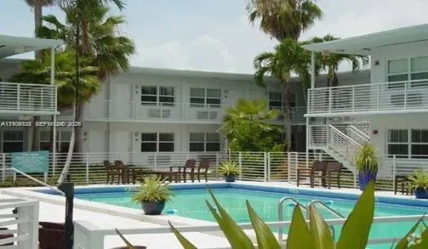 In-ground swimming pool with a white concrete deck, enclosed by a white metal perimeter fence, and surrounded by mature palm trees