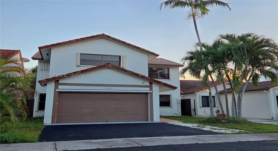 Two-story exterior featuring a light-toned stucco façade, terracotta-style roof tiles, an attached garage with a sectional door, and mature palm trees