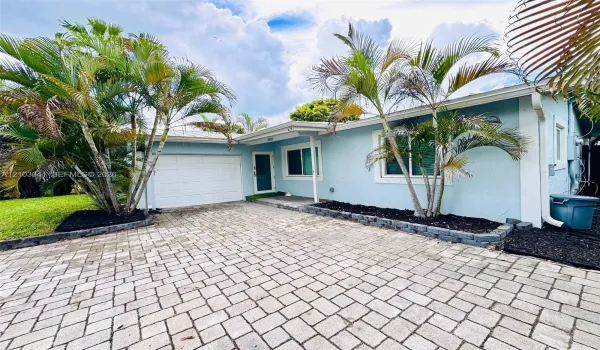 Single-story exterior with light blue stucco siding, paver driveway, single-car garage, and integrated landscaping featuring mature palms and stone-bordered garden beds