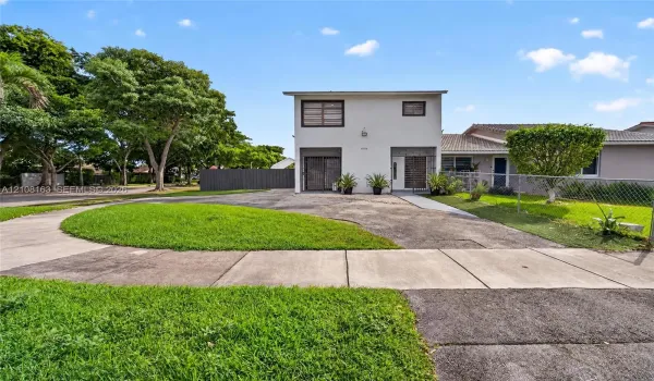Two-story exterior with a light-toned stucco facade, paved driveway, concrete walkway, and chain-link perimeter fencing