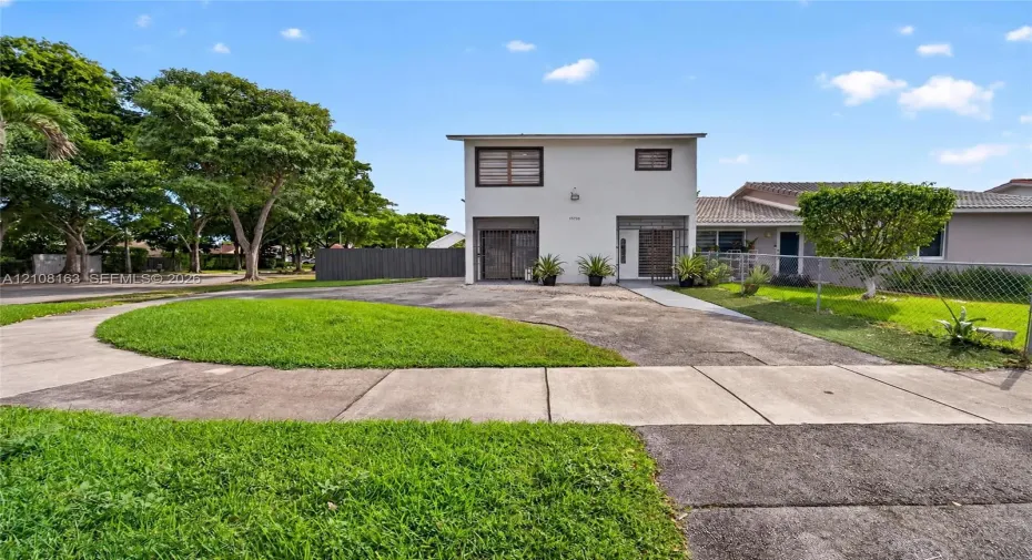 Two-story exterior with a light-toned stucco facade, paved driveway, concrete walkway, and chain-link perimeter fencing