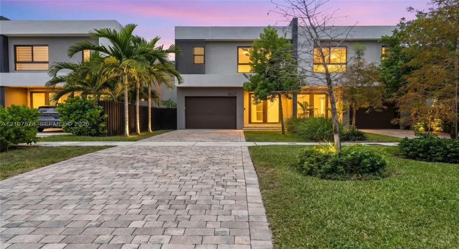 Modern architectural exterior featuring a concrete facade, paver driveway, integrated garage, and floor-to-ceiling glass entry doors
