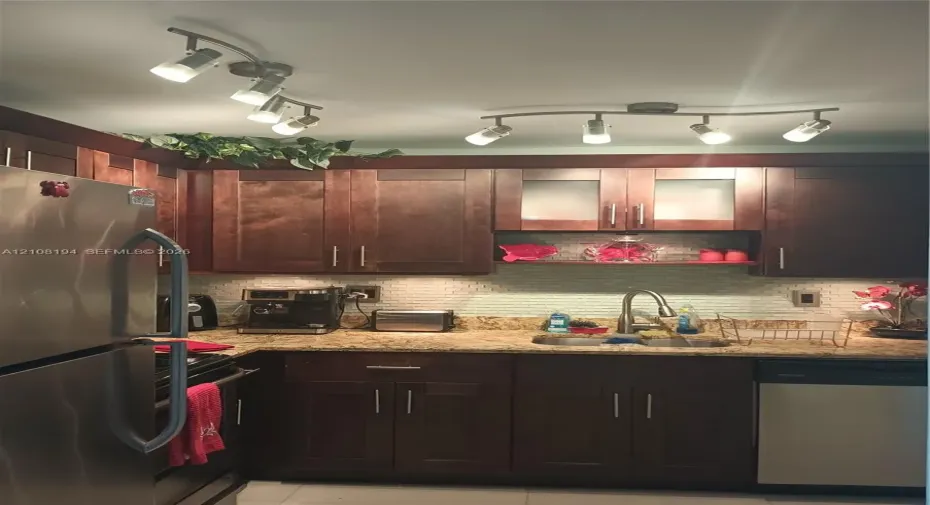 Dark wood cabinetry featuring brushed metal hardware, stone-finish countertops, textured tile backsplash, stainless steel appliances, and dual adjustable track lighting fixtures