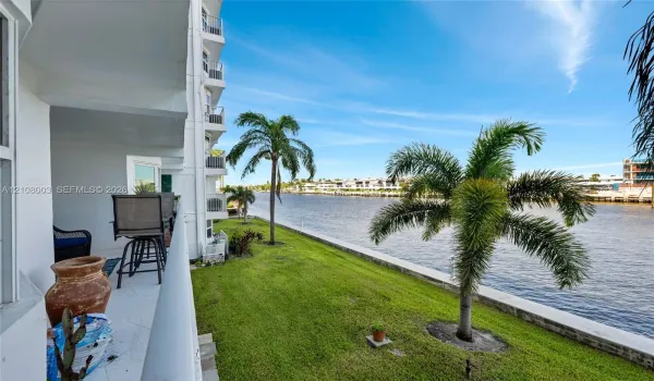 Covered balcony featuring white tiled flooring and a metal railing, overlooking a waterfront lawn with palm trees and a concrete bulkhead