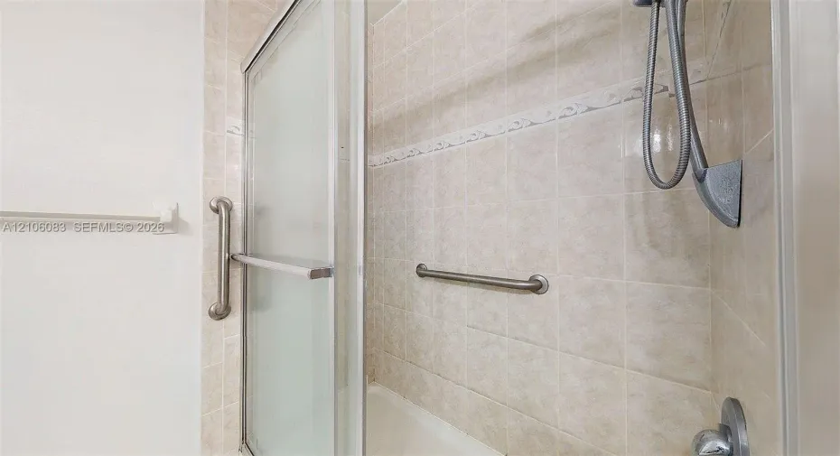 Shower area featuring tiled walls with decorative accents, sliding glass door, handheld shower head, and wall-mounted metal grab bars
