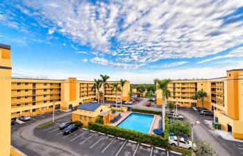 Multi-story residential complex featuring an outdoor swimming pool, paved surface parking, and mature palm trees