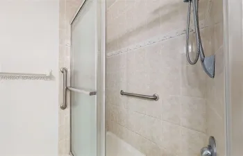 Shower area featuring tiled walls with decorative accents, sliding glass door, handheld shower head, and wall-mounted metal grab bars