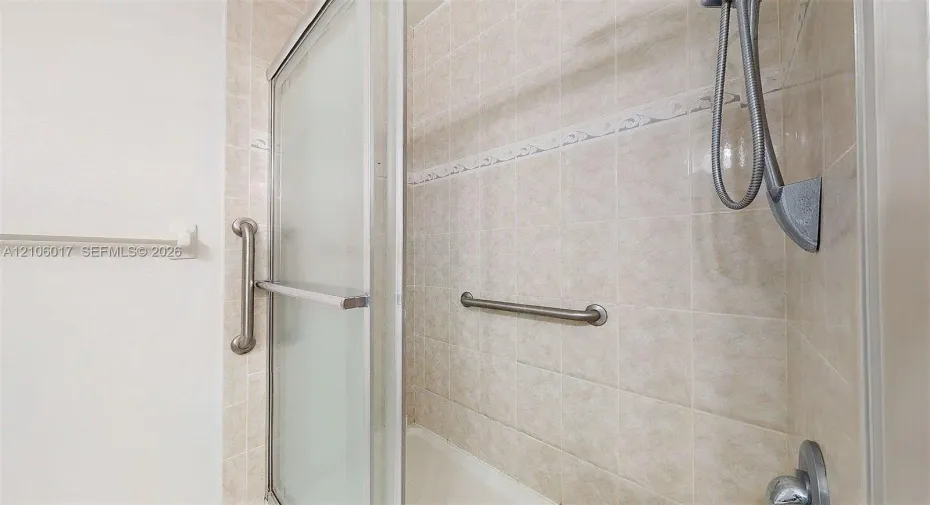 Shower area featuring tiled walls with decorative accents, sliding glass door, handheld shower head, and wall-mounted metal grab bars
