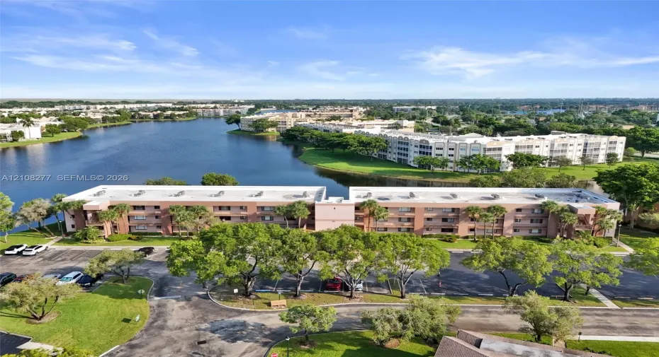 Multi-story residential building featuring stucco exterior, expansive lake frontage, mature trees, and dedicated surface parking