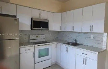 Kitchen featuring white flat-panel cabinetry, stone-patterned countertops, coordinating backsplash, stainless steel microwave, and stainless steel refrigerator