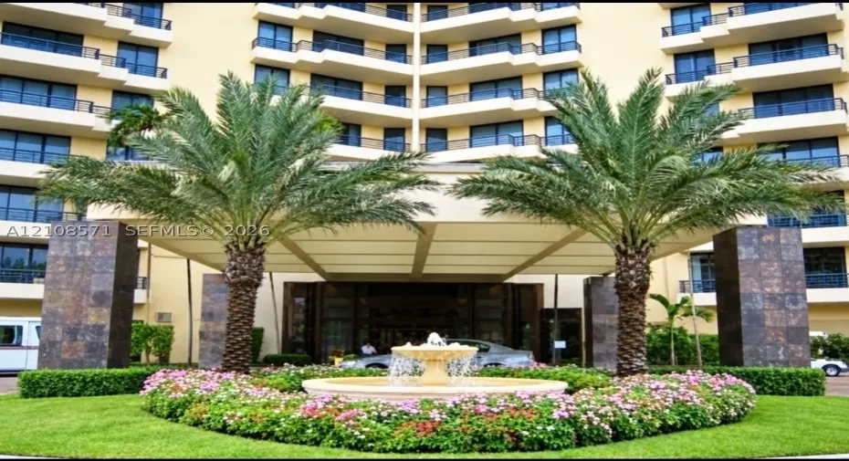 Multi-story building facade featuring a covered porte-cochère, decorative central stone fountain, mature palm trees, and manicured landscaping with flowering perennials