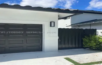 Stucco exterior featuring a dark-finish paneled garage door, scalloped privacy gate, and modern rectangular wall-mounted light fixture under a tiled roofline