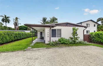 Single-story exterior featuring a covered front porch, light-colored stucco finish, stone-surfaced driveway, and mature perimeter hedging