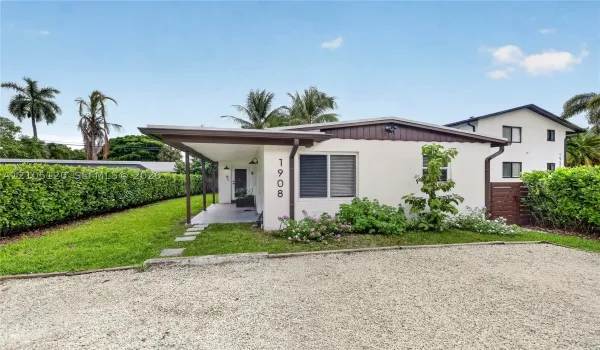 Single-story exterior featuring a covered front porch, light-colored stucco finish, stone-surfaced driveway, and mature perimeter hedging