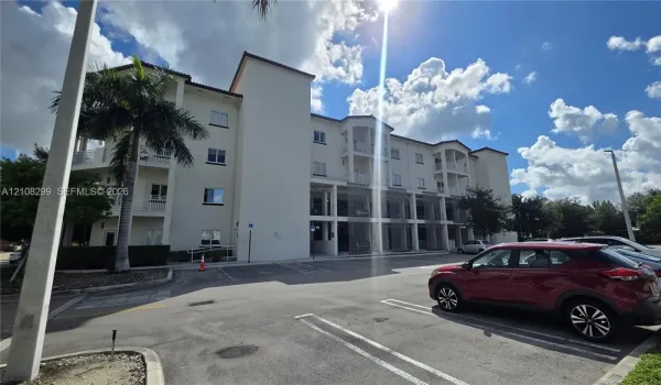 Multi-story exterior featuring a light-toned stucco façade, tile roof detailing, recessed balconies, and paved surface parking