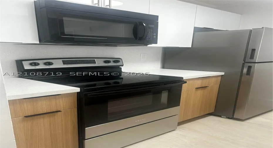 Kitchen featuring light wood-finish cabinetry, white stone-composite countertops, stainless steel refrigerator, black electric range, and over-the-range microwave