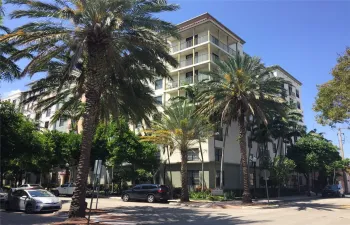 Mid-rise multi-story building exterior featuring a stucco facade, tiered balconies with glass railings, a flat roofline, and mature palm tree landscaping