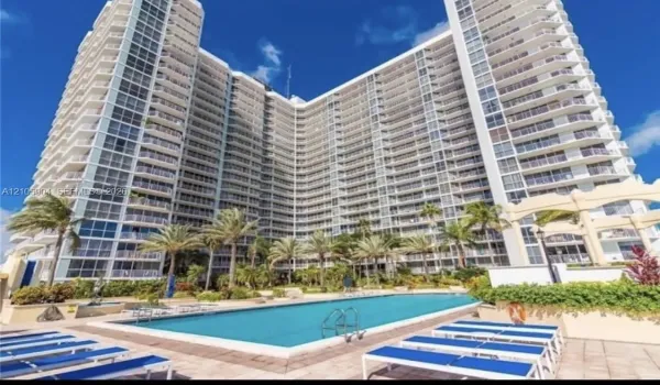 High-rise building exterior featuring a multi-story facade with wraparound balconies, accompanied by an outdoor swimming pool, paved sun deck, and mature palm trees
