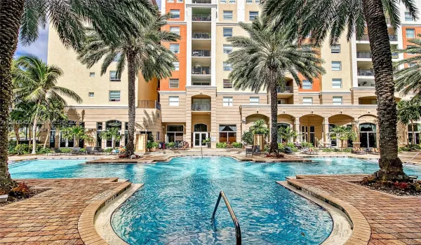 Resort-style swimming pool featuring a brick-paver deck, arched architectural detailing, mature palm trees, and multi-story residential facade