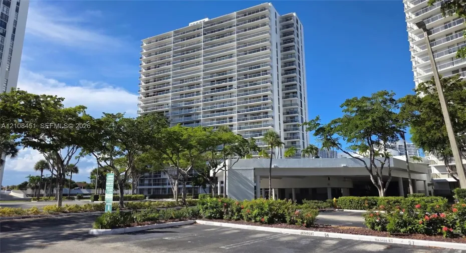 High-rise residential building featuring expansive private balconies, a multi-level concrete structure, and ground-level covered parking, complemented by mature landscaping