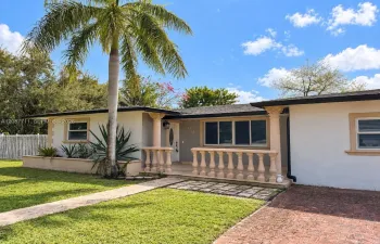 Stucco exterior featuring a covered front porch with decorative balustrade and columns, brick-paved driveway, and mature landscaping