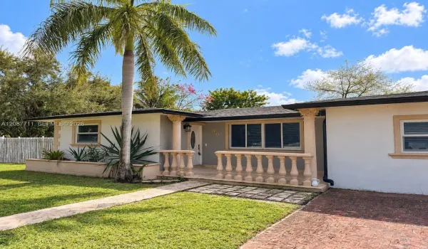 Stucco exterior featuring a covered front porch with decorative balustrade and columns, brick-paved driveway, and mature landscaping