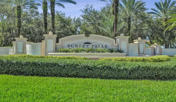 Stucco community signage featuring decorative masonry pillars, integrated lighting fixtures, tiered architectural detailing, and mature palm landscaping
